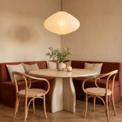 Comedor pequeño con banco en esquina de terciopelo terracota, mesa redonda de travertino y lámpara de papel encendida