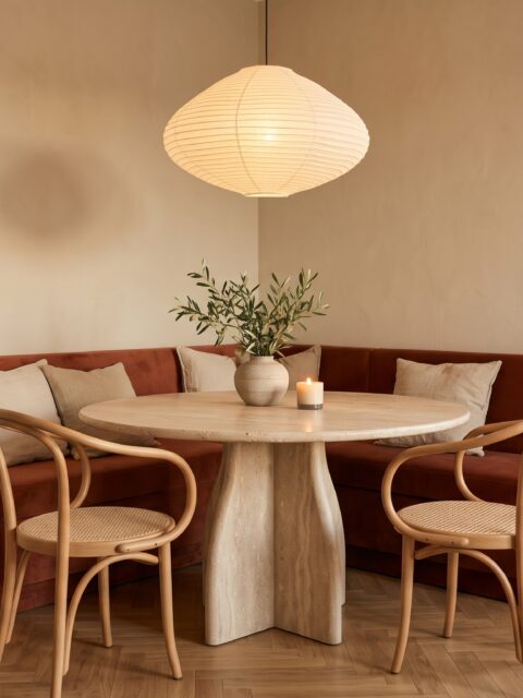 Comedor pequeño con banco en esquina de terciopelo terracota, mesa redonda de travertino y lámpara de papel encendida